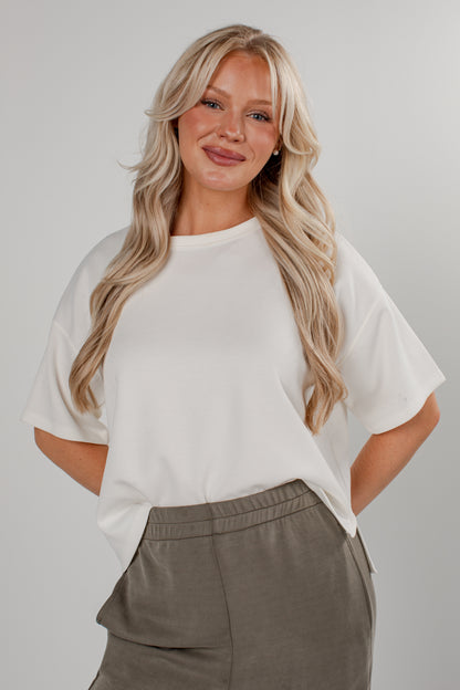 A blonde woman wearing a white short-sleeve crewneck top and gray pants, standing against a light gray studio background.