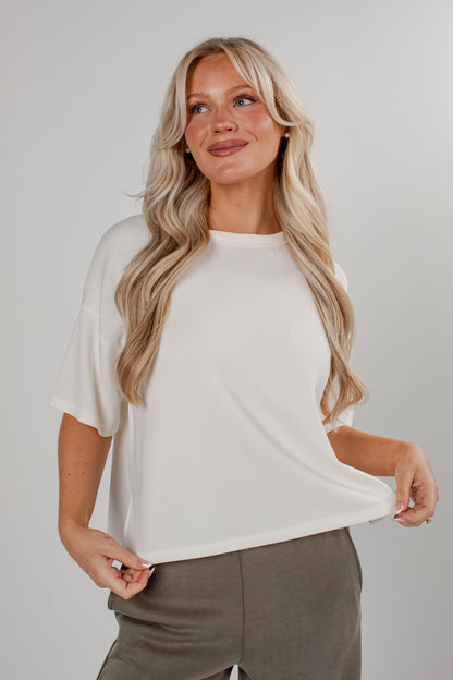 A blonde woman wearing a short-sleeve off-white knit top and gray trousers, posing against a light gray studio background.