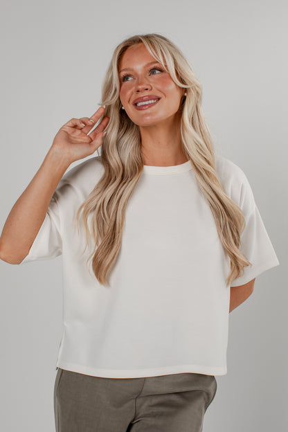 A woman with long blonde hair wearing an oversized off-white short-sleeve crop top and gray pants, posing against a neutral light gray background.
