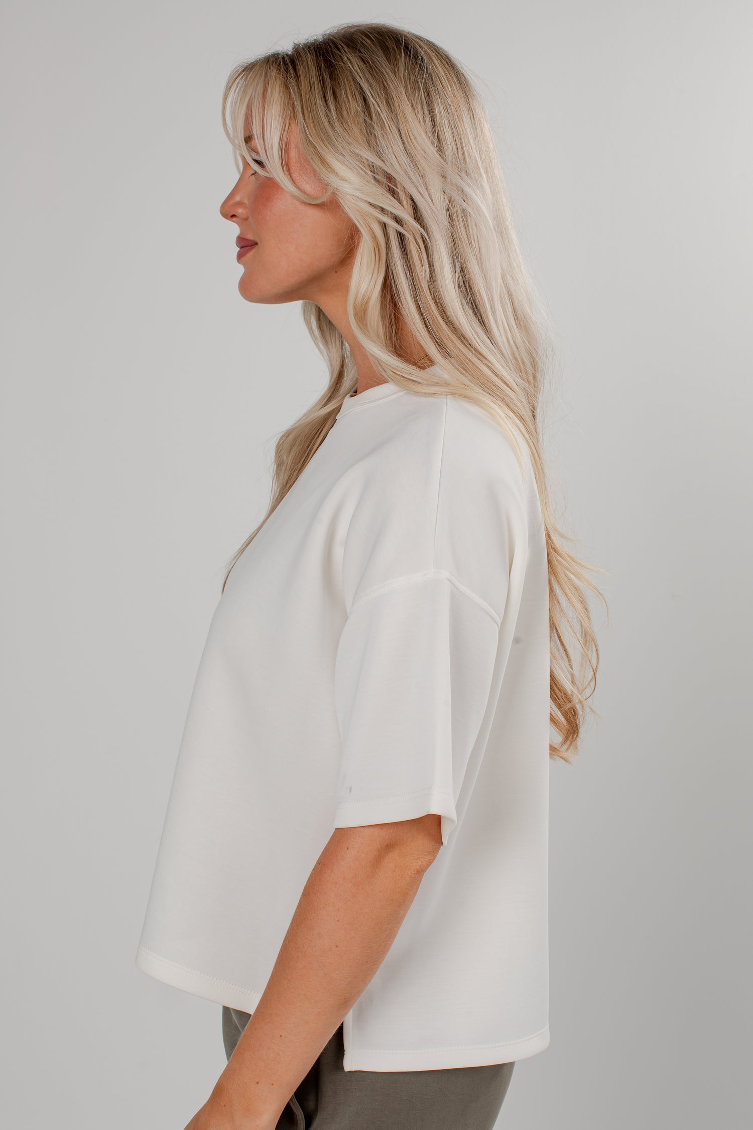 A woman with blonde hair is shown in profile wearing a loose-fitting white short-sleeve top against a plain light gray studio background.