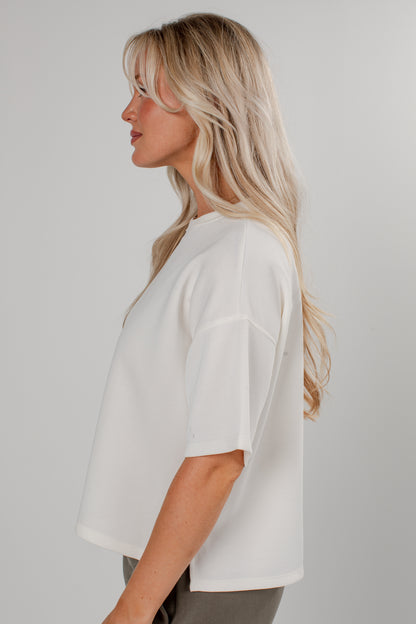 A woman with blonde hair is shown in profile wearing a loose-fitting white short-sleeve top against a plain light gray studio background.