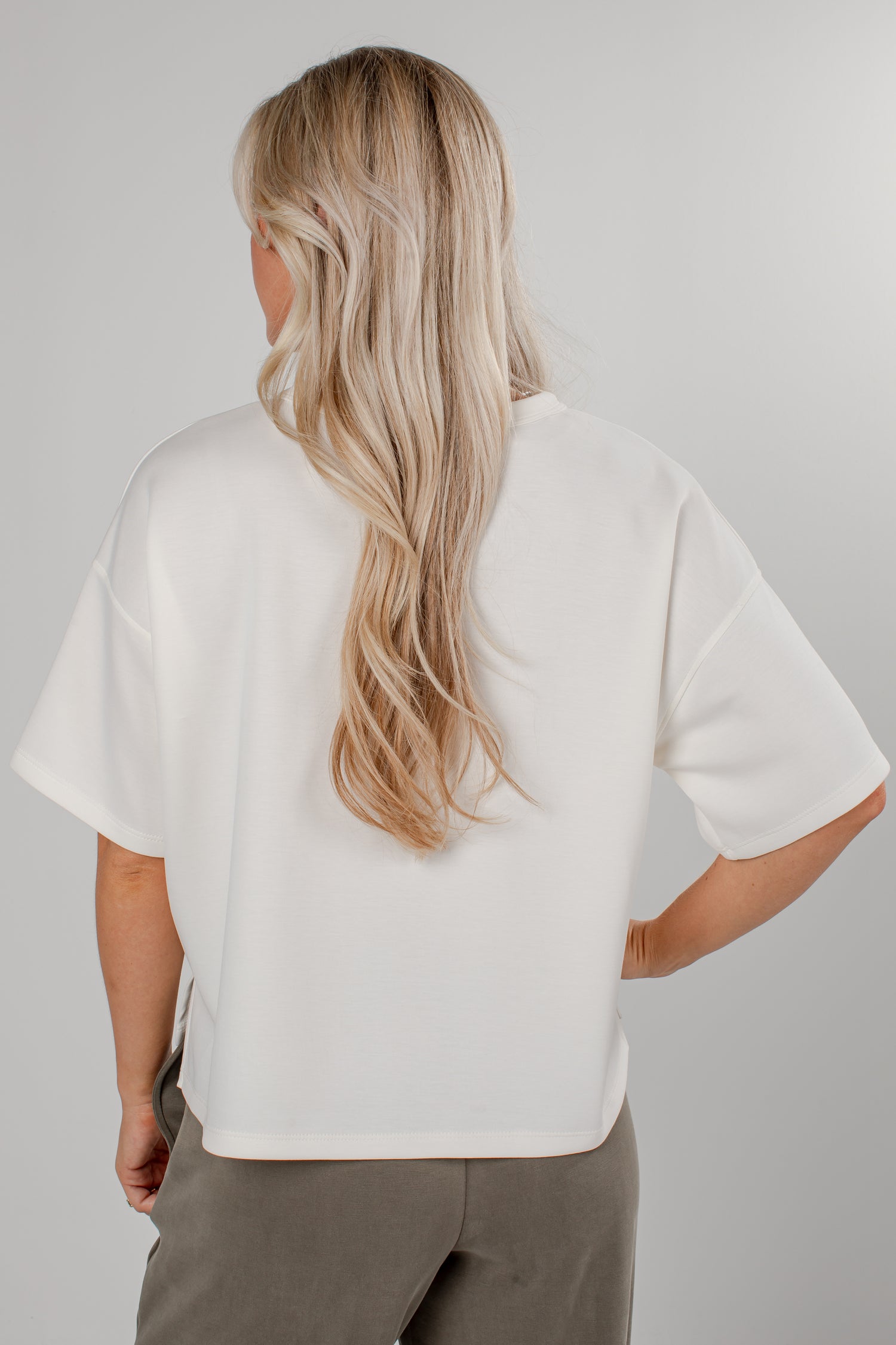 A woman with long blonde hair is shown from the back wearing an oversized white short-sleeve top and taupe pants against a light gray studio background.