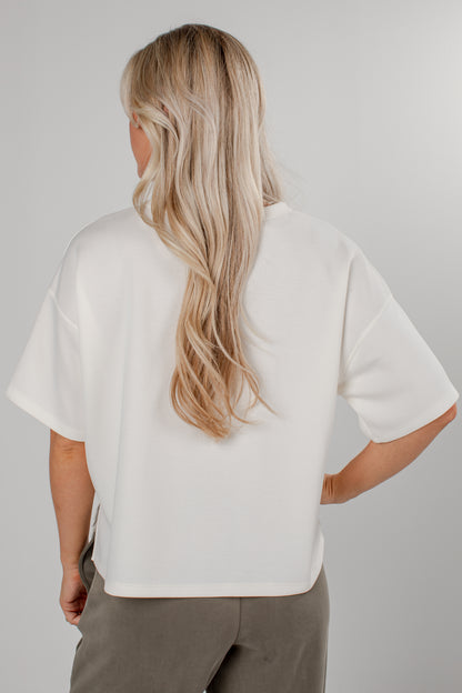 A woman with long blonde hair is shown from the back wearing an oversized white short-sleeve top and taupe pants against a light gray studio background.