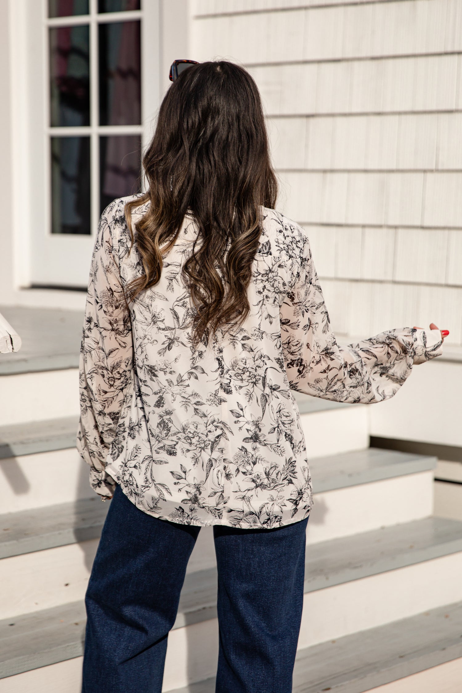 Woman standing on a set of stairs wearing a floral blouse and dark pants.