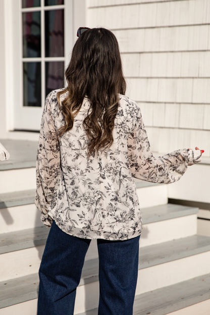 Woman standing on a set of stairs wearing a floral blouse and dark pants.