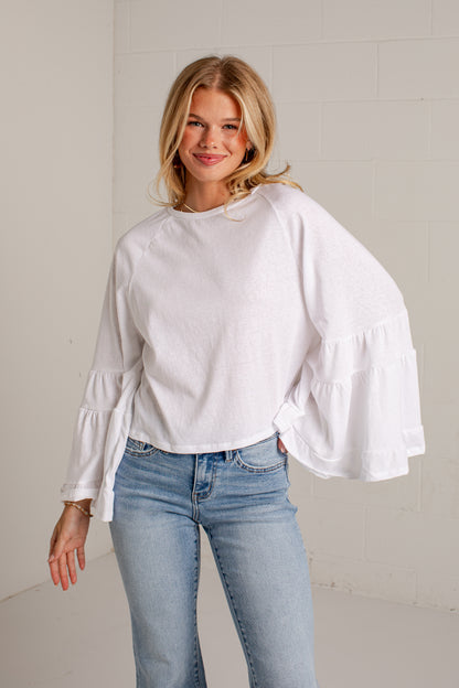 A woman wearing a white long-sleeve blouse with tiered ruffle sleeves and light blue jeans, standing against a white brick wall.