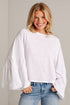 A smiling blonde woman wearing a white long-sleeve top with tiered ruffle sleeves and light blue jeans, standing against a light gray brick wall.