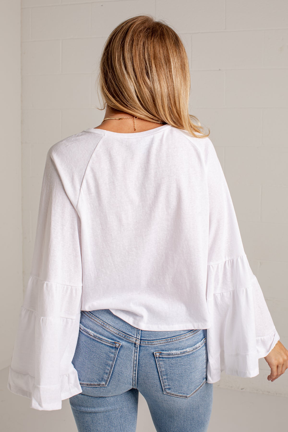 A woman viewed from behind wearing a white long-sleeve top with layered ruffle cuffs and light blue jeans, standing against a light gray brick wall.