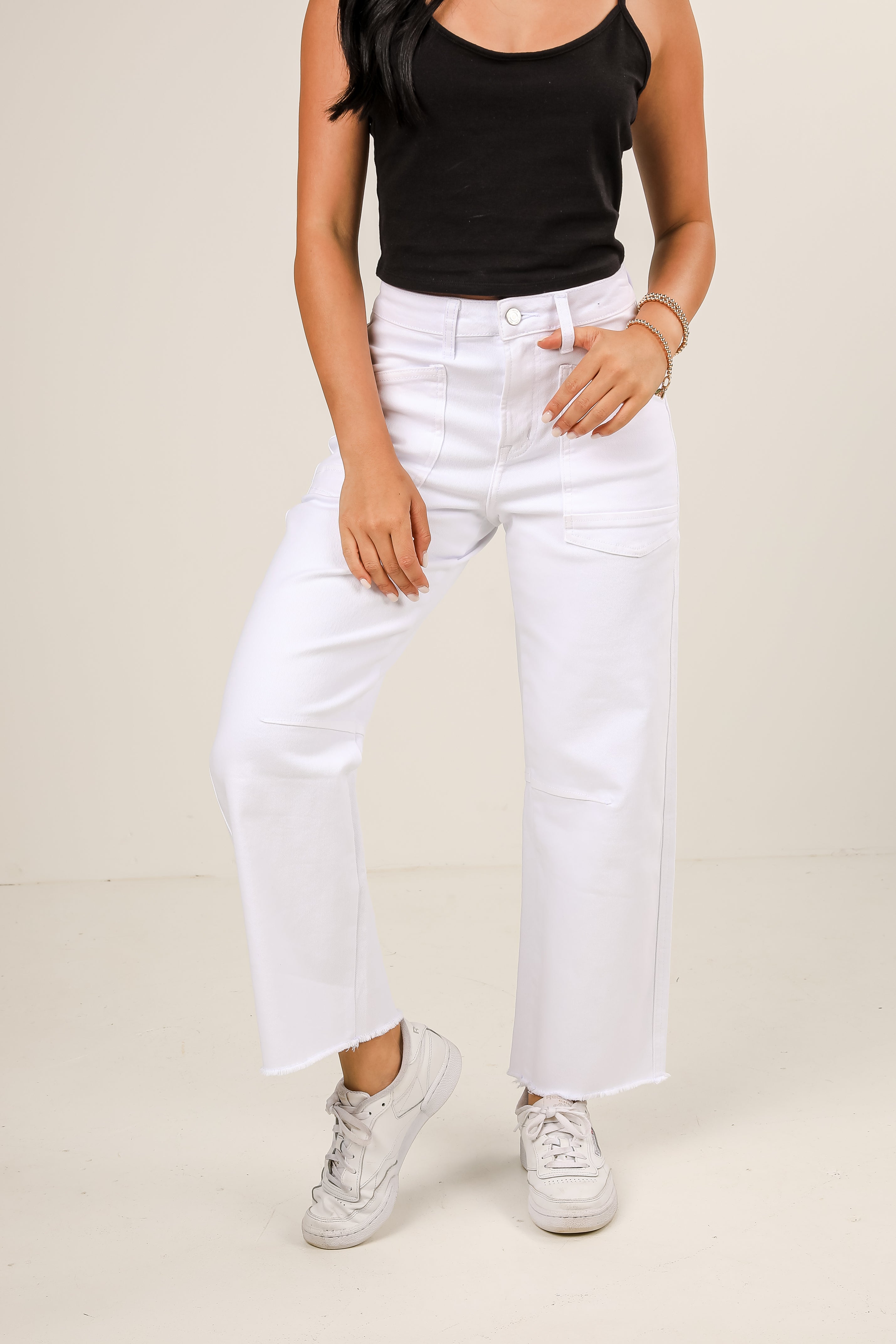 Person wearing a black tank top, white jeans, and white sneakers on a plain background