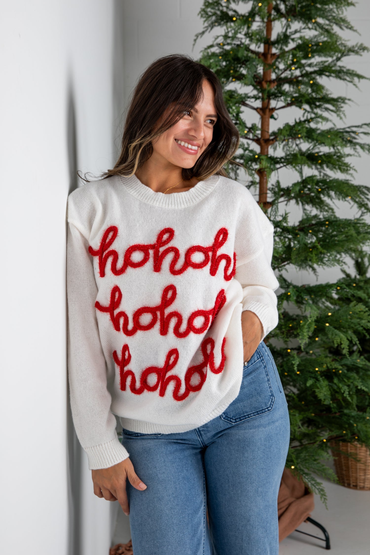 Woman wearing a white sweater with red text standing next to a Christmas tree.