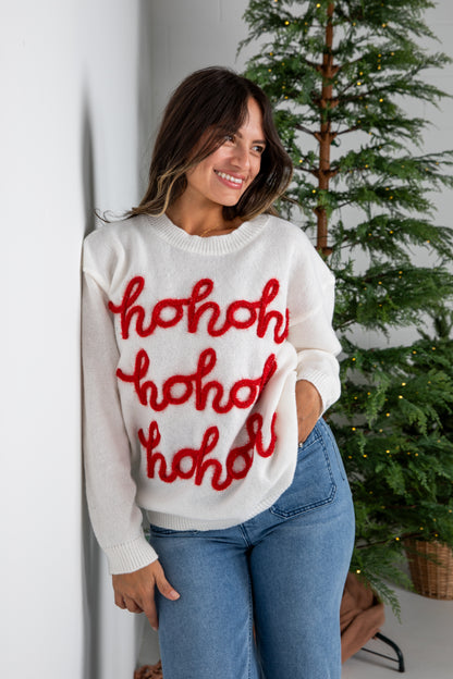 Woman wearing a white sweater with red text standing next to a Christmas tree.