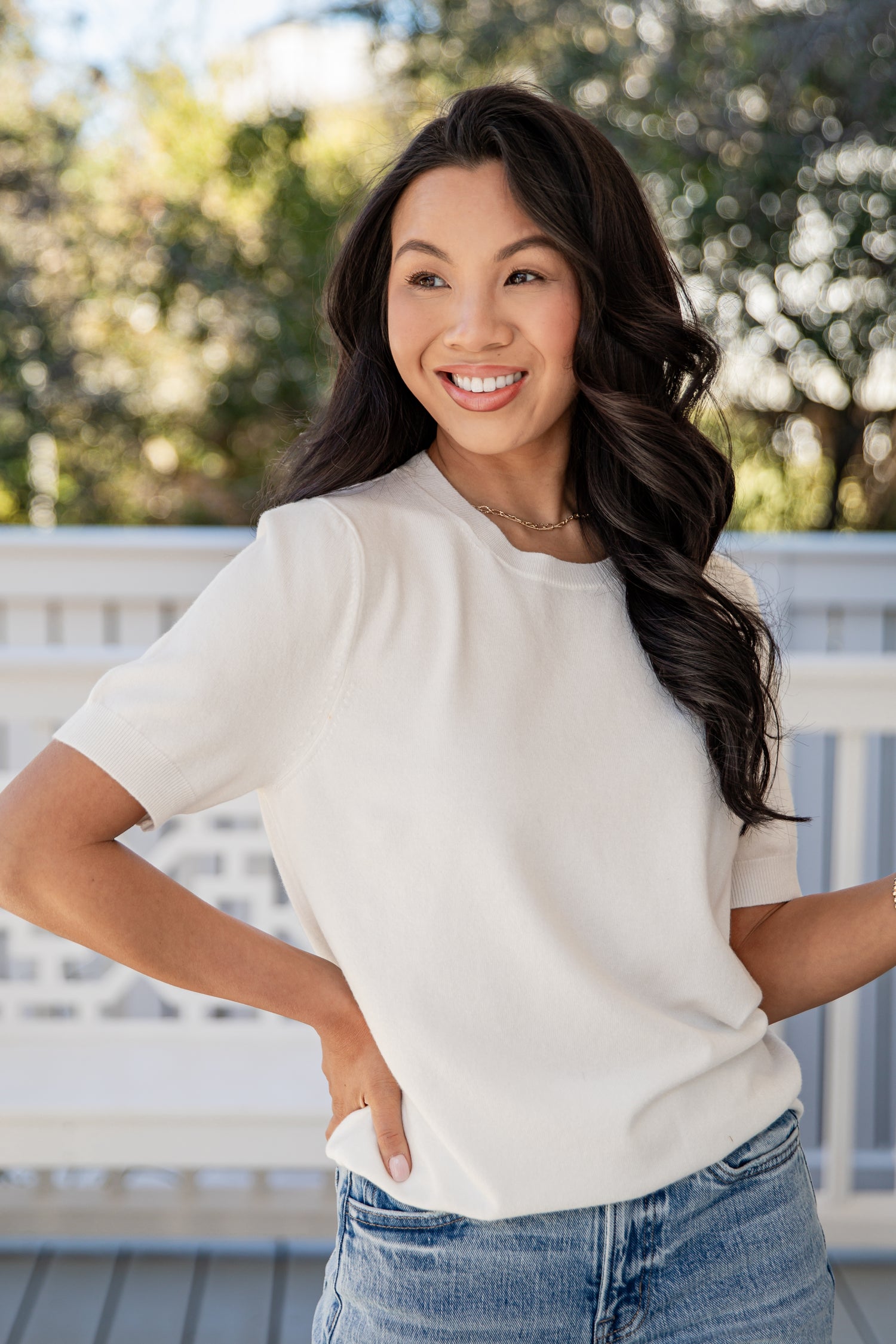 Woman wearing a white t-shirt and blue jeans standing outdoors with greenery in the background