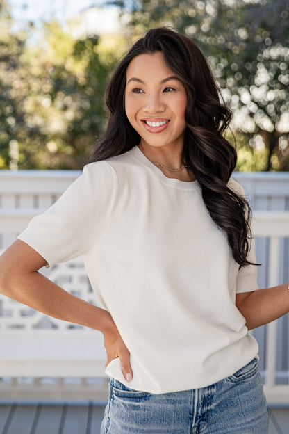 Woman wearing a white t-shirt and blue jeans standing outdoors with greenery in the background