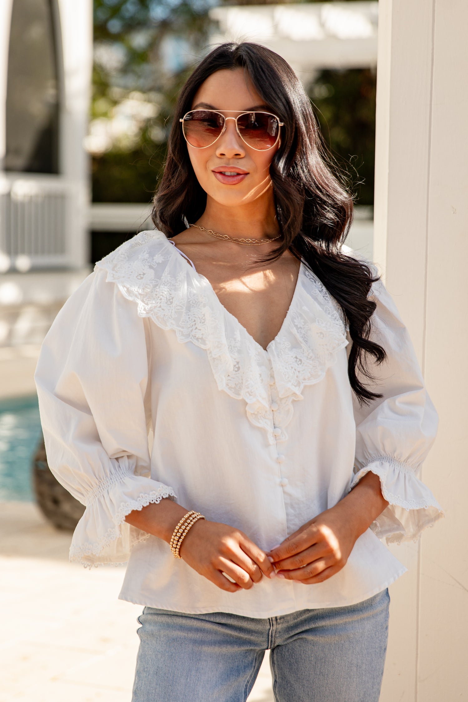 Woman wearing a white blouse and sunglasses by a poolside.