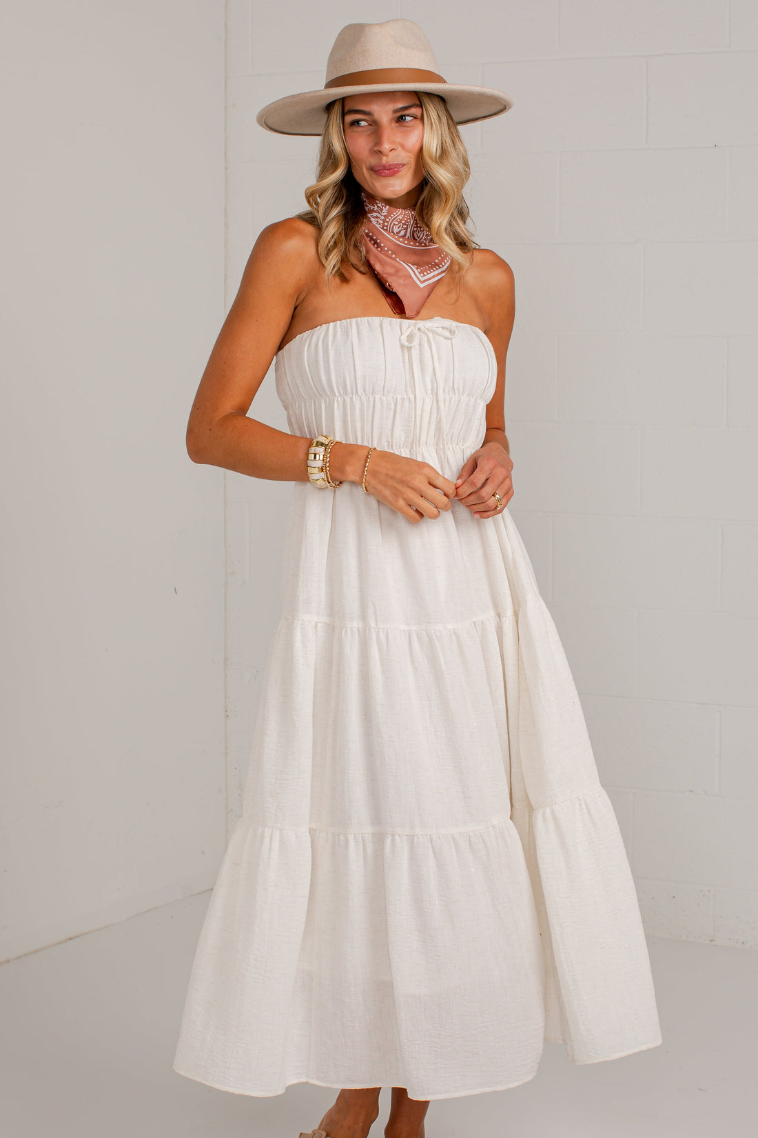 A woman modeling a strapless tiered midi dress in off-white, styled with a beige wide-brim hat, a pink patterned neck scarf, and beige flat shoes against a white studio background.