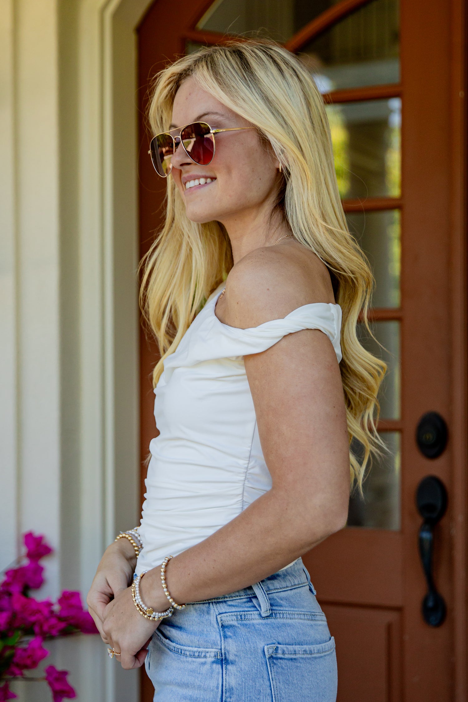 A blonde woman wearing a white off-the-shoulder top, light blue jeans, and round sunglasses stands in front of a brown wooden door.