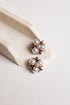 A pair of gold-toned flower-shaped earrings with white faux pearls, displayed on a cream-colored ceramic stand against a light off-white background.