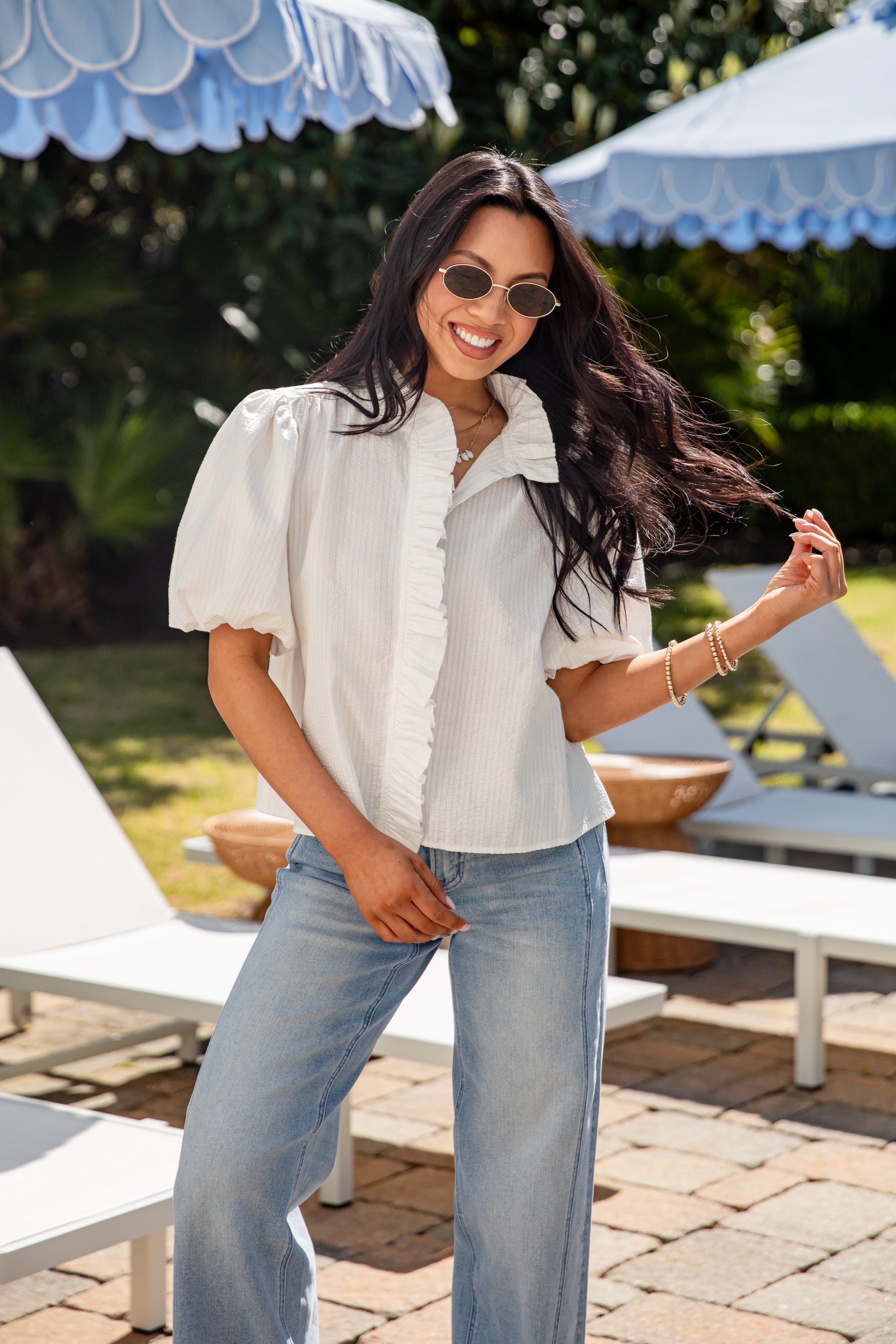 Woman in a white blouse and blue jeans standing outdoors with lounge chairs and umbrellas in the background.