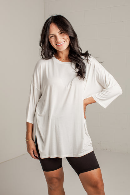 A woman modeling a white oversized long-sleeve tunic top with black fitted shorts against a light gray studio wall.