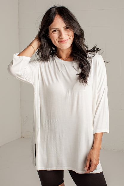 A woman with long dark hair wearing a loose-fitting white long-sleeve tunic top and black leggings, standing against a light gray wall.