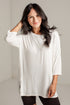 A woman with long dark hair wearing a loose-fitting white long-sleeve tunic top and black leggings, standing against a light gray wall.