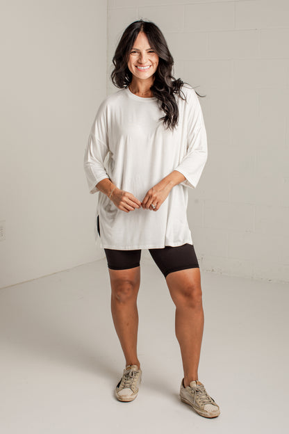 A woman modeling a loose-fitting off-white three-quarter sleeve tunic top, paired with black bike shorts and beige sneakers, standing in a bright, neutral studio setting.