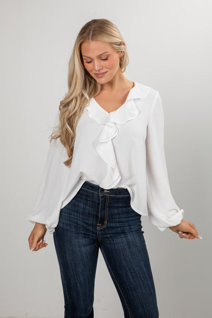 Woman wearing a white blouse with ruffle details and dark jeans on a plain background