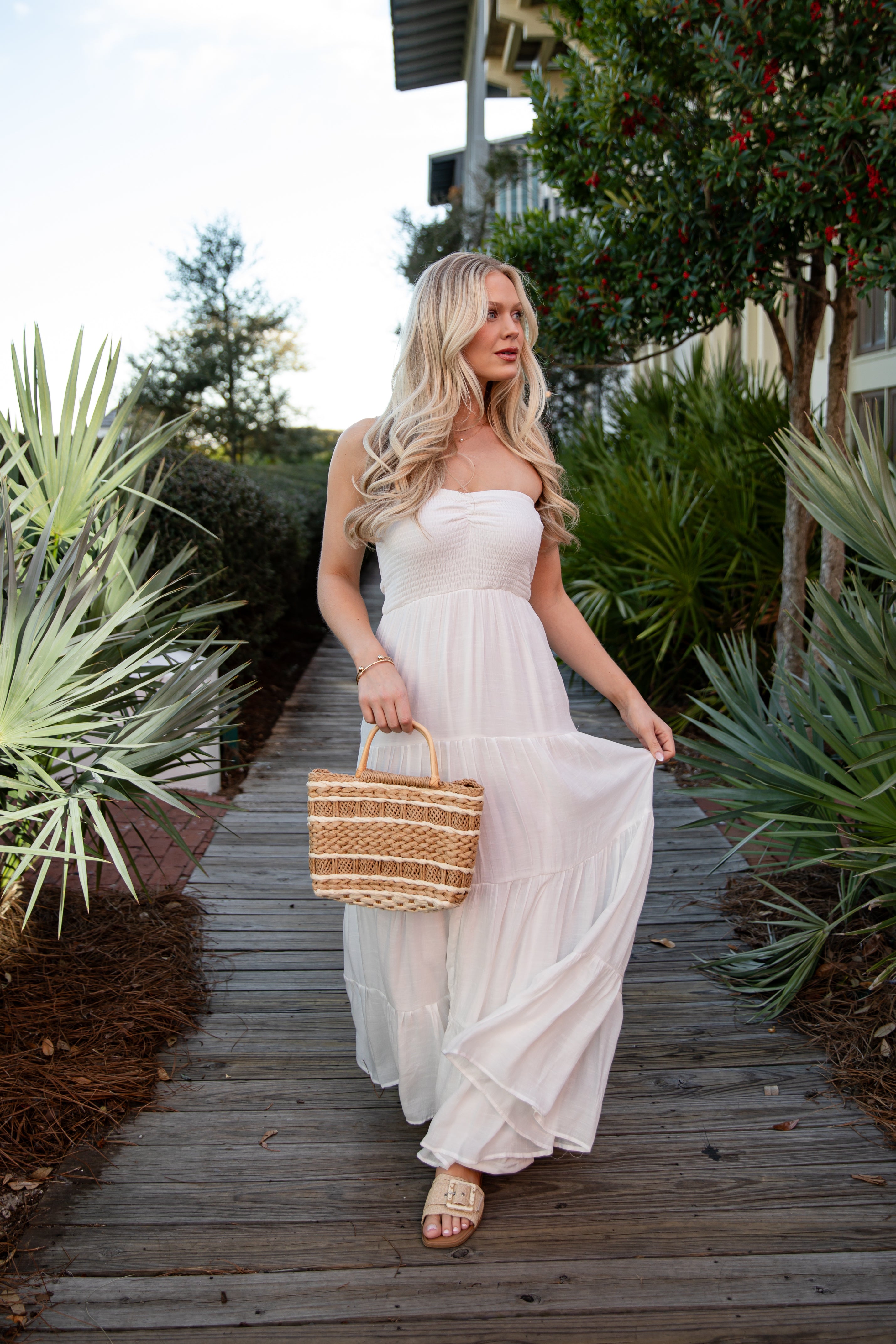 Woman in a white dress holding a straw bag on a wooden path with greenery.