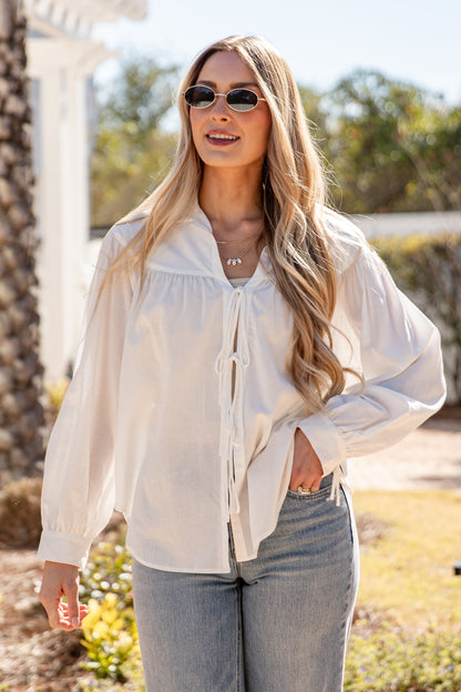 Woman wearing a white blouse and sunglasses outdoors with greenery in the background