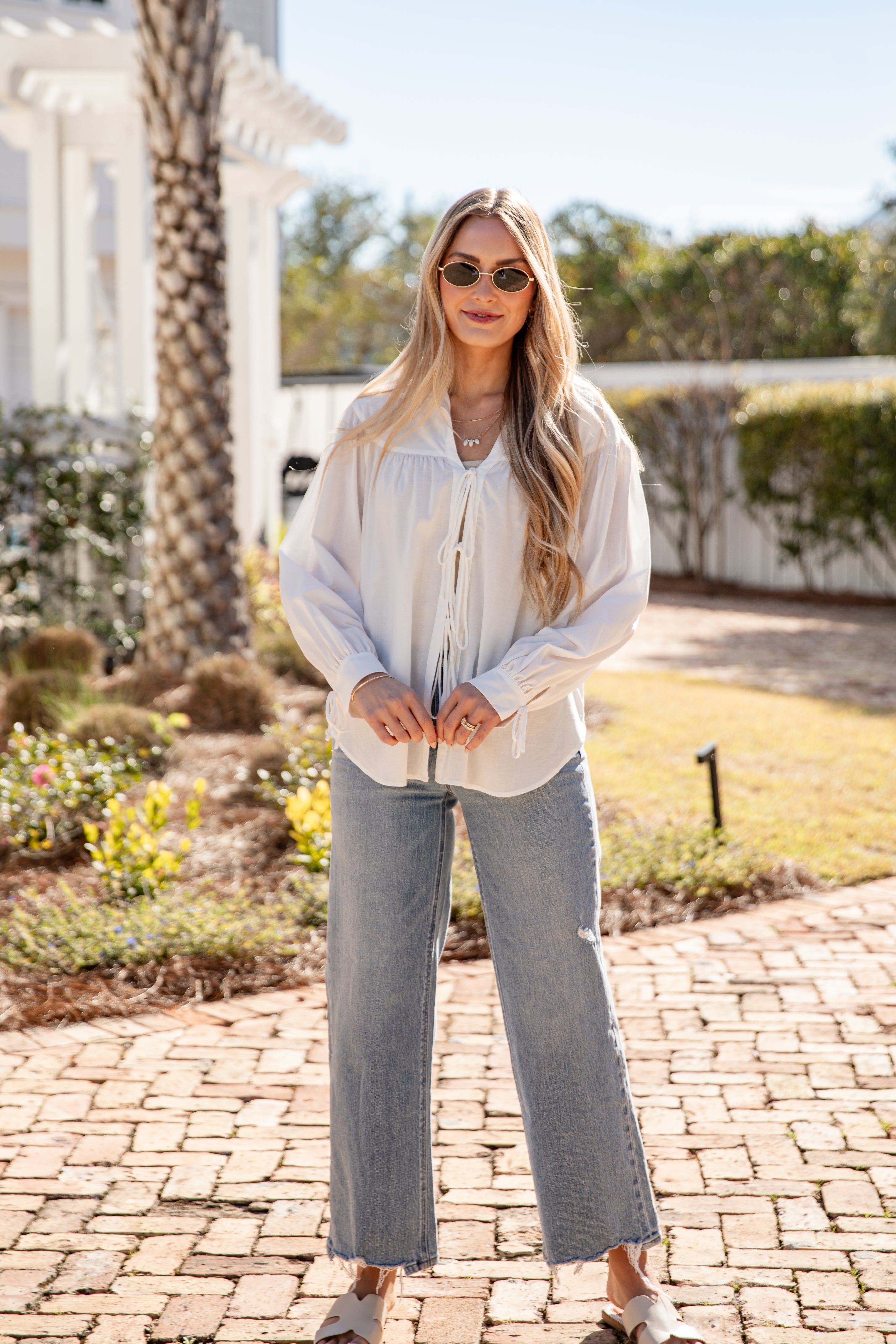 Woman wearing a light-colored blouse and jeans standing on a brick path with greenery in the background