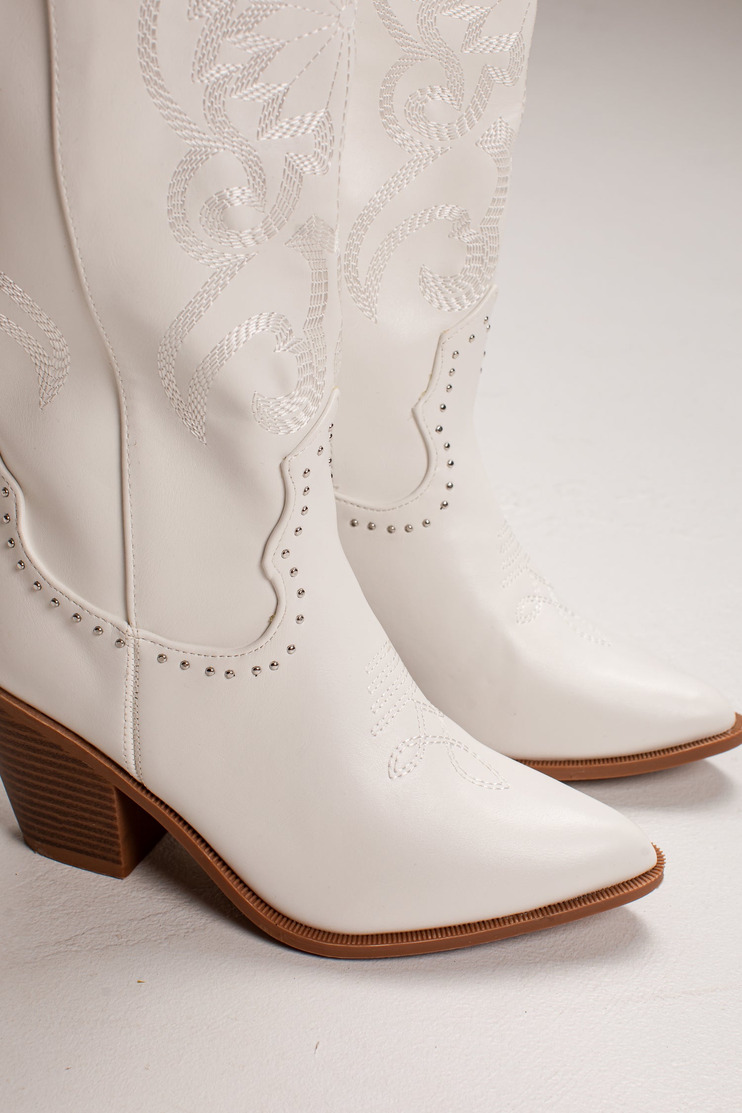 A pair of white faux leather cowboy boots with decorative western embroidery, studded trim, and brown stacked block heels, shown against a light gray studio background.