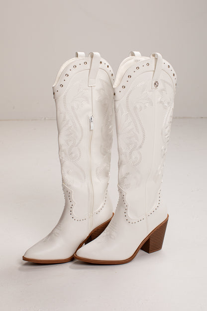A pair of white leather cowboy boots with decorative stitching, silver studs, and brown stacked block heels, shown against a plain light gray studio background.