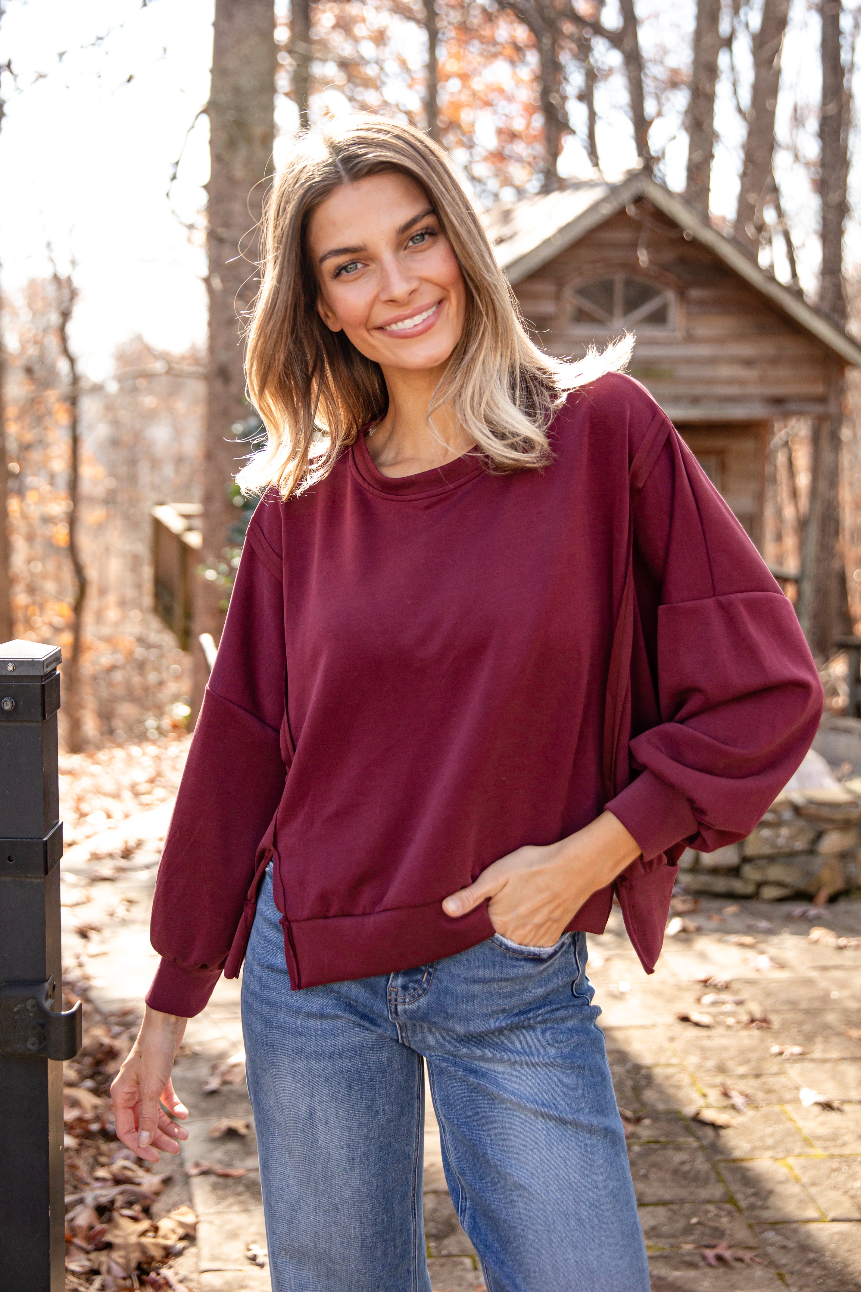 Woman wearing a maroon sweatshirt and blue jeans standing outdoors with trees and a wooden structure in the background.