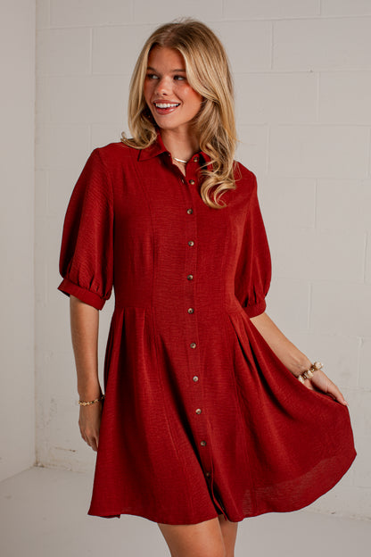 A woman wearing a red button-front shirt dress with puff sleeves and a flared skirt, standing against a white painted brick wall.