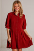 A smiling blonde woman models a short red button-up shirt dress with puff sleeves and a flared skirt, standing against a white painted brick wall.