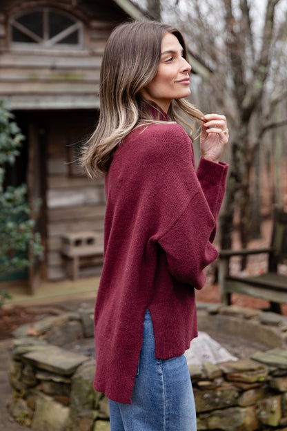 Woman wearing a maroon sweater and blue jeans standing outdoors.