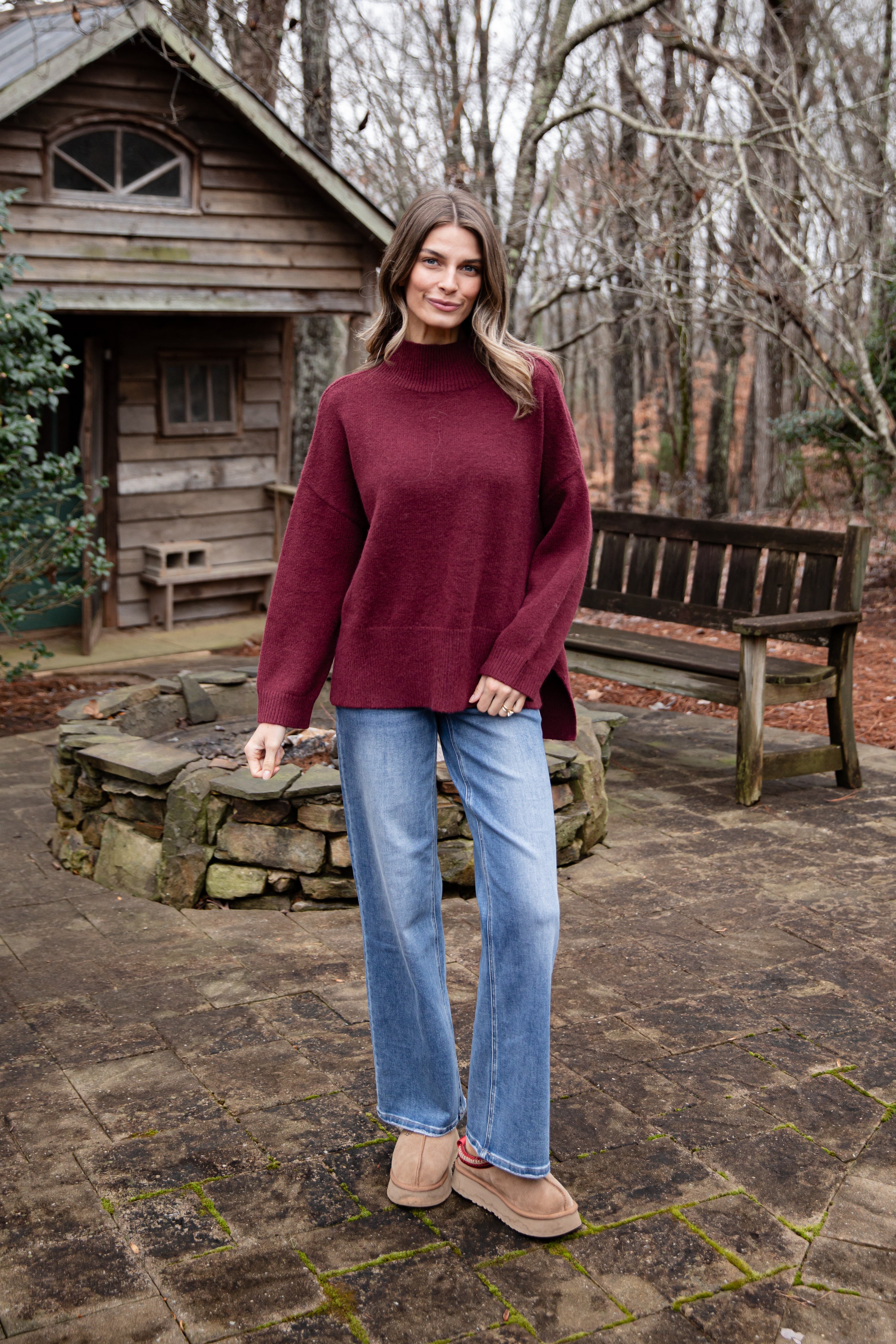 Woman wearing a burgundy sweater and blue jeans standing in front of a wooden cabin.