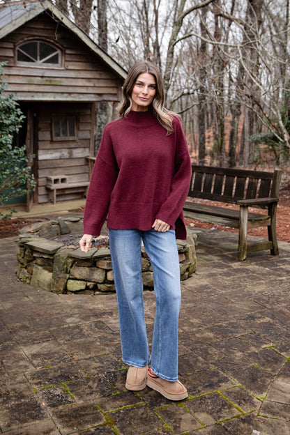 Woman wearing a burgundy sweater and blue jeans standing in front of a wooden cabin.