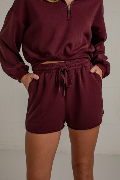 A person wearing a matching burgundy lounge set consisting of a cropped half-zip long-sleeve top and drawstring shorts, standing against a light gray brick wall.
