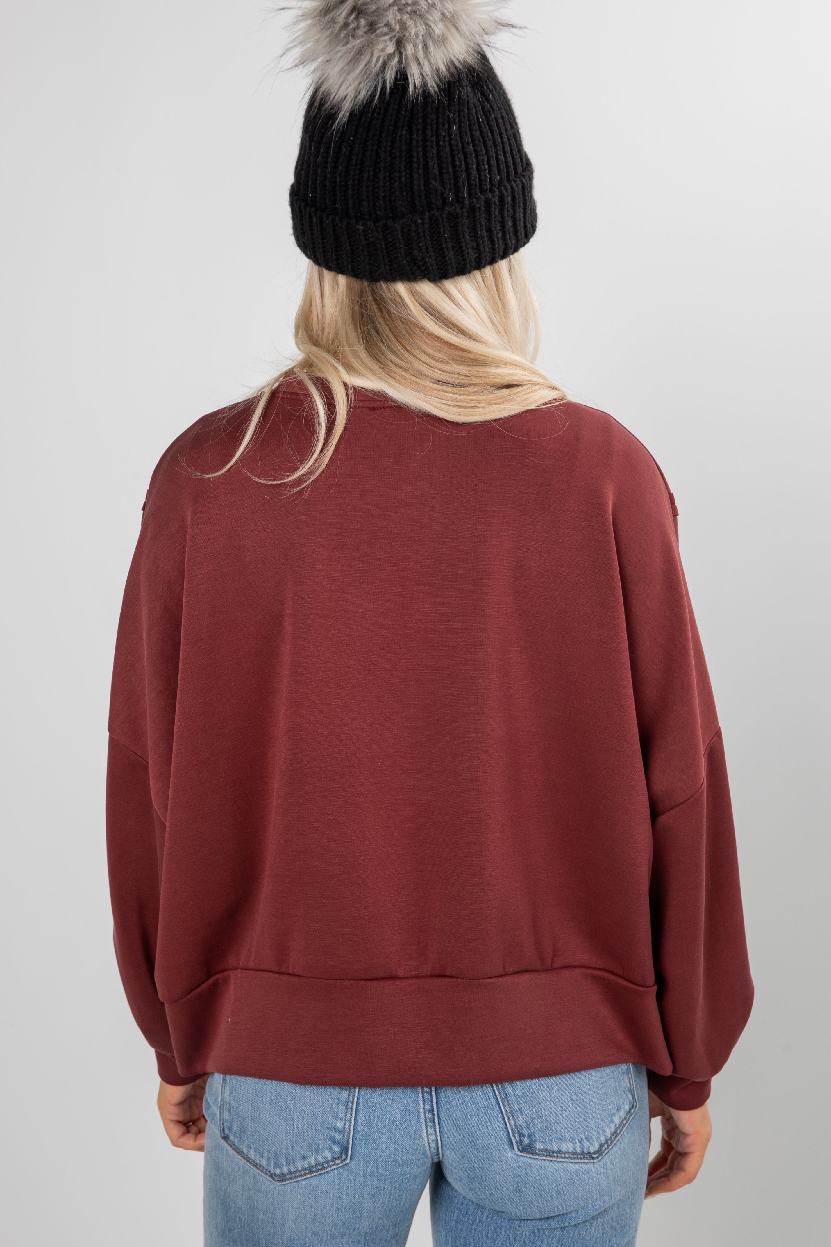 Person wearing a maroon sweatshirt, black knit beanie with fur, and blue jeans on a plain background