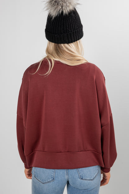 Person wearing a maroon sweatshirt, black knit beanie with fur, and blue jeans on a plain background