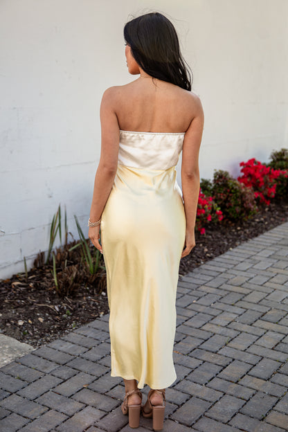 Woman in a strapless white dress standing on a paved path with a white wall and red flowers in the background.
