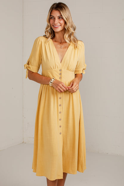 A woman modeling a mustard yellow midi dress with short sleeves, a V-neckline, and a button-down front, standing against a plain off-white studio wall.