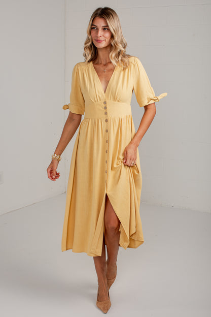 A woman modeling a mustard yellow midi dress with short flutter sleeves, a V-neckline, button-front detail, and a high-low hem, photographed in a bright white studio setting.