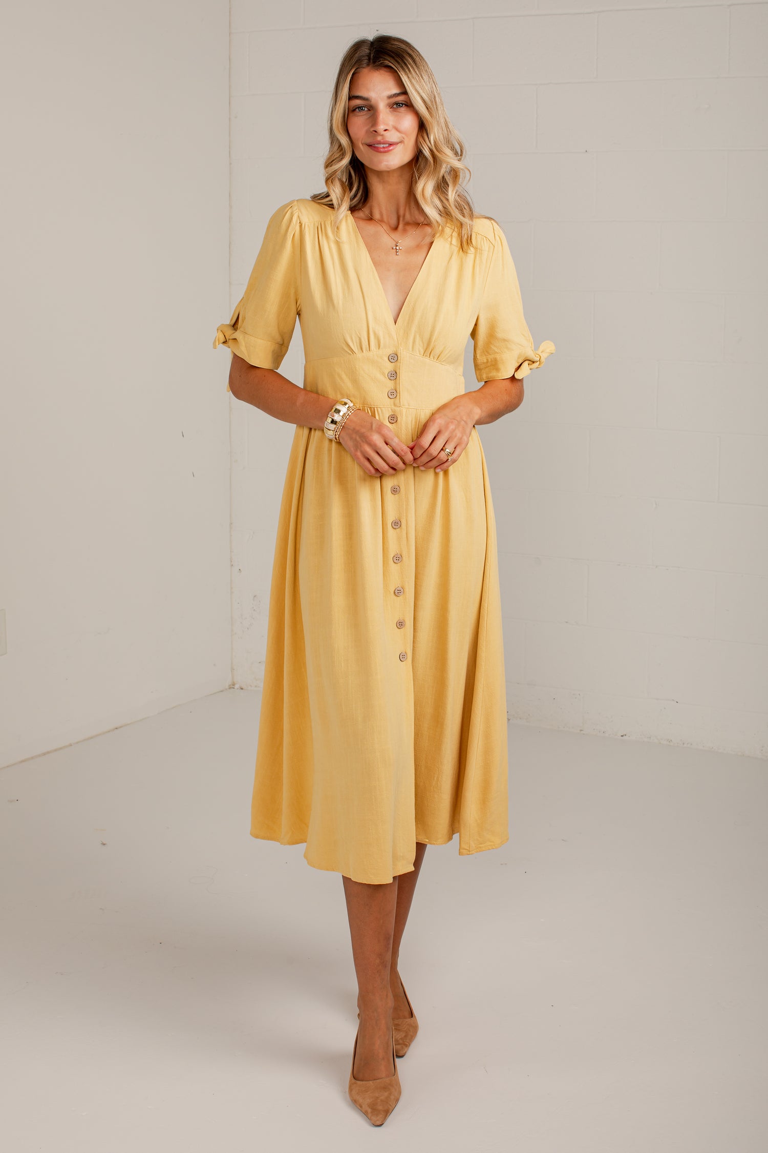 A woman modeling a yellow midi dress with short flutter sleeves, a V-neckline, and a button-front detail, standing in a bright studio setting.