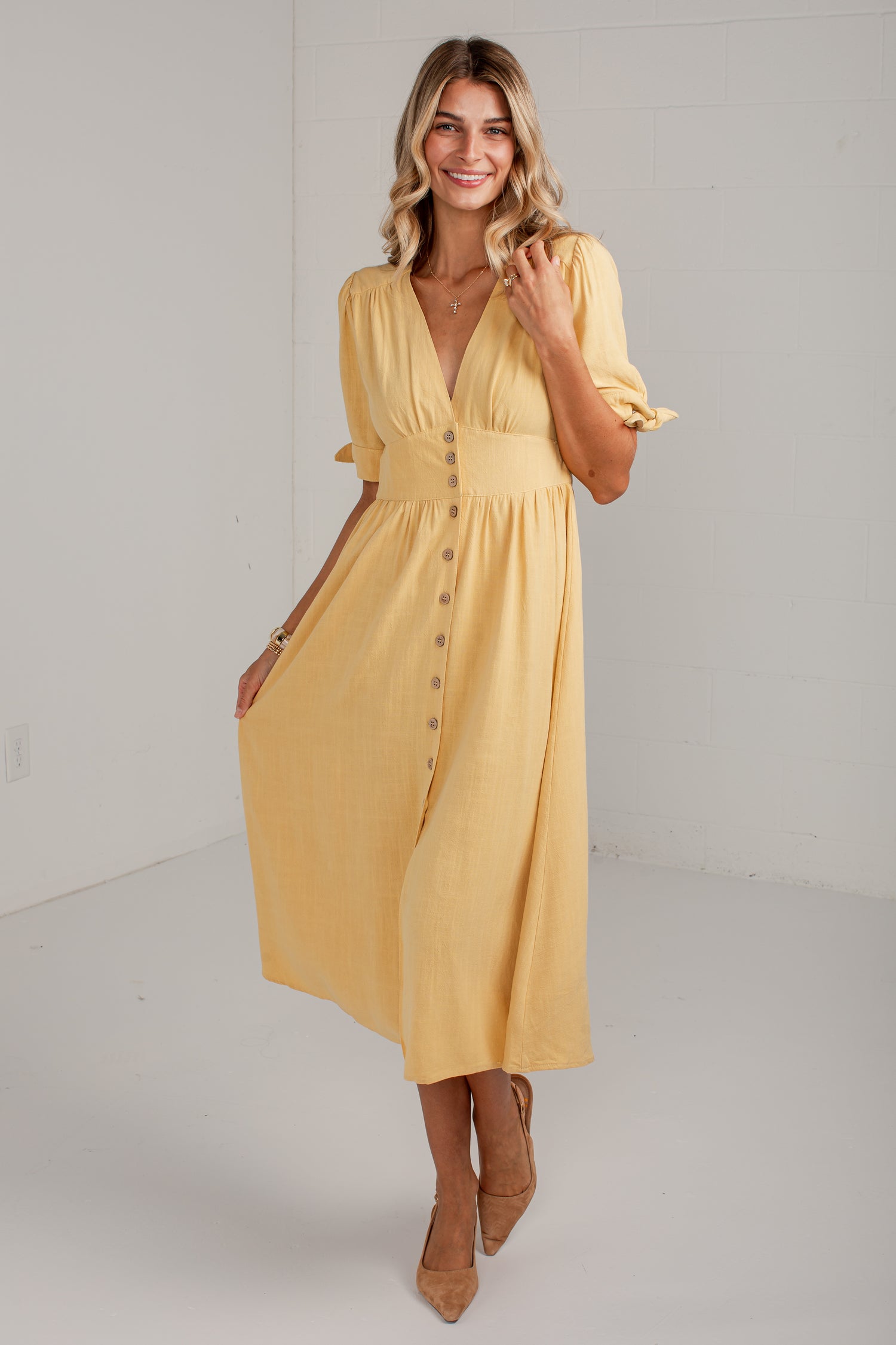 A woman modeling a yellow midi dress with puff sleeves, a V-neckline, and a button-down front, standing in a bright white studio setting.