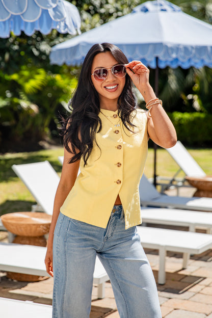 Woman in yellow shirt and blue jeans standing outdoors under a blue umbrella.