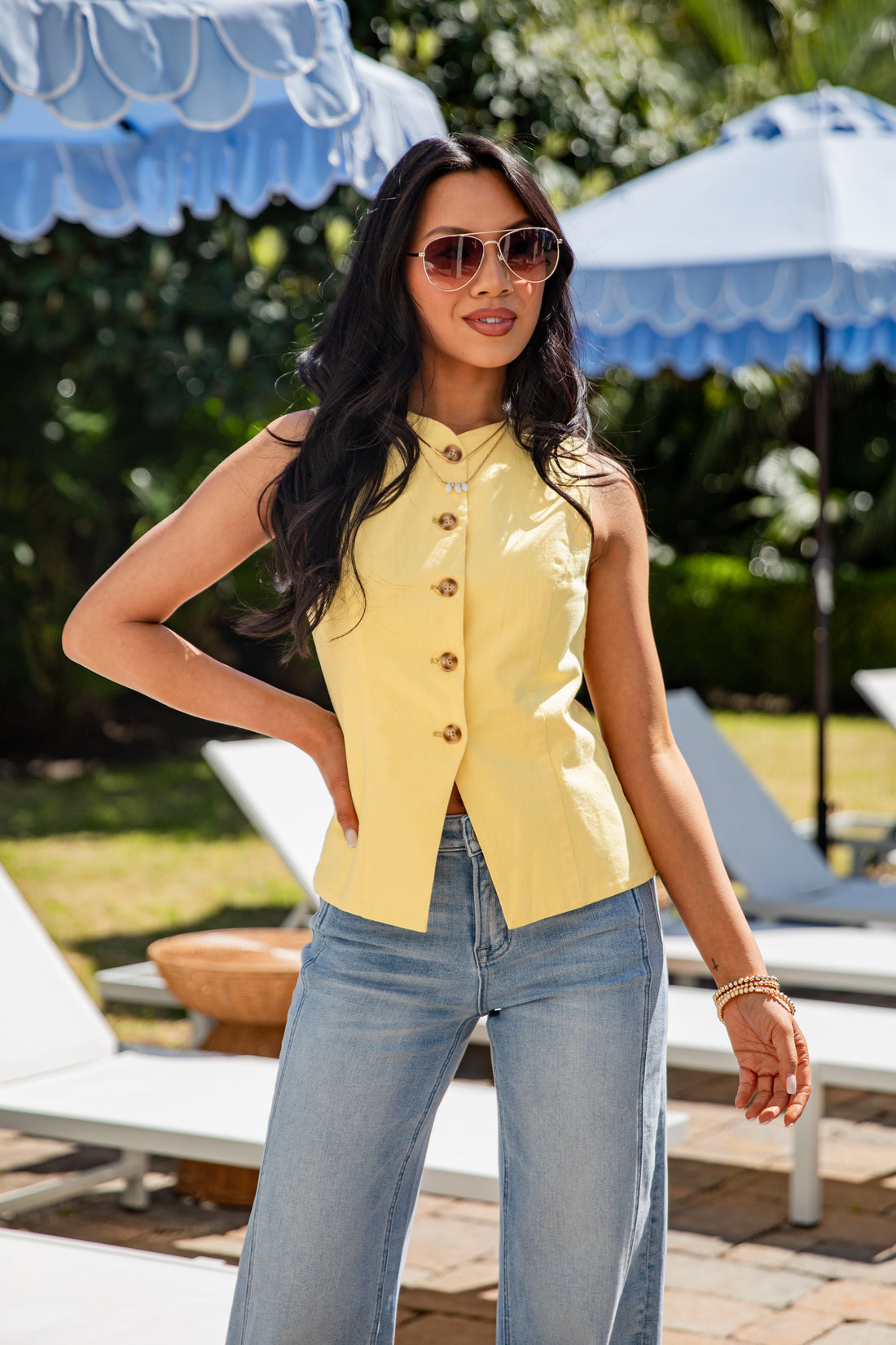 Woman wearing a yellow sleeveless top and blue jeans standing by lounge chairs with blue umbrellas in the background.