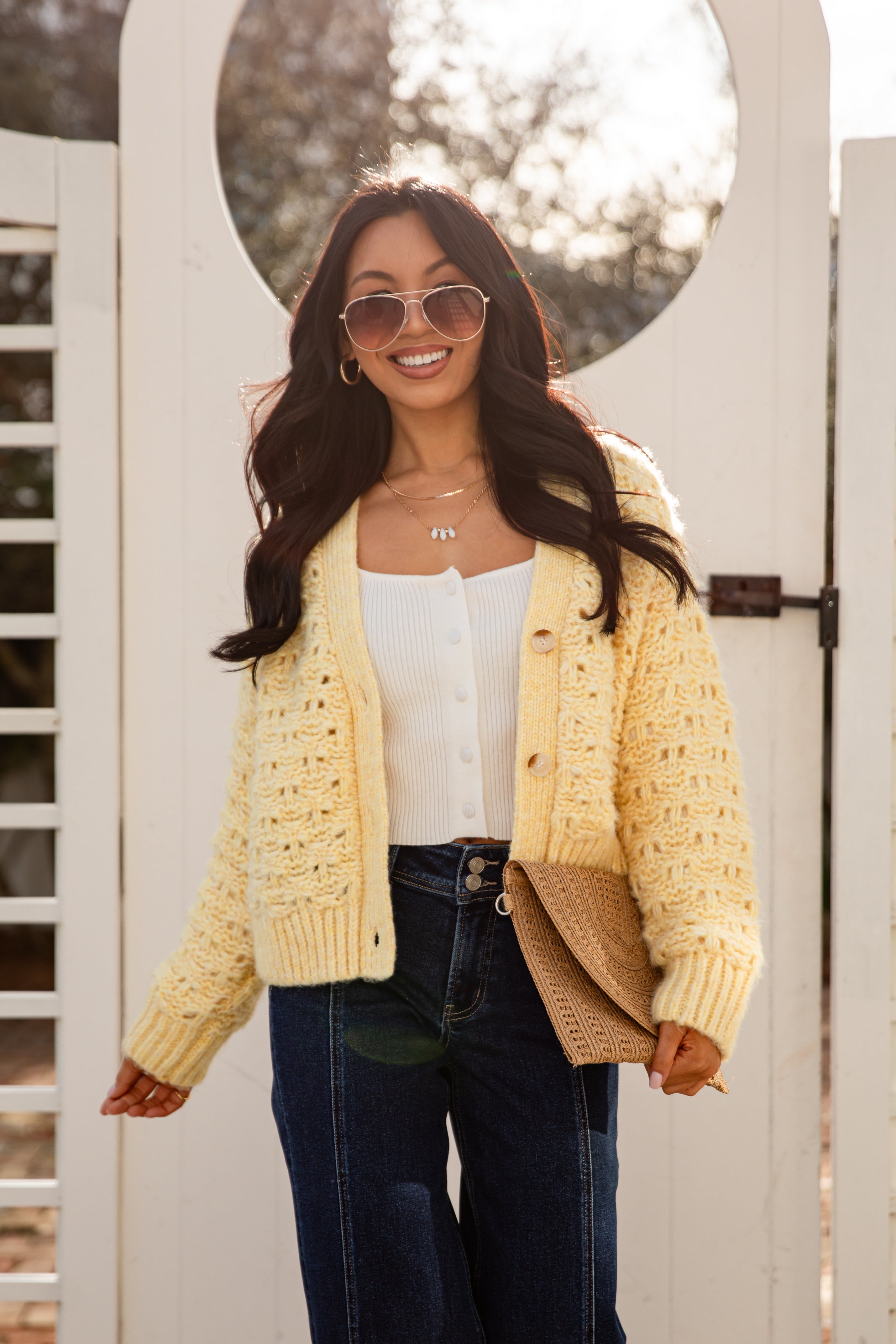 Woman wearing a yellow cardigan, white top, and dark jeans standing in front of a white gate.