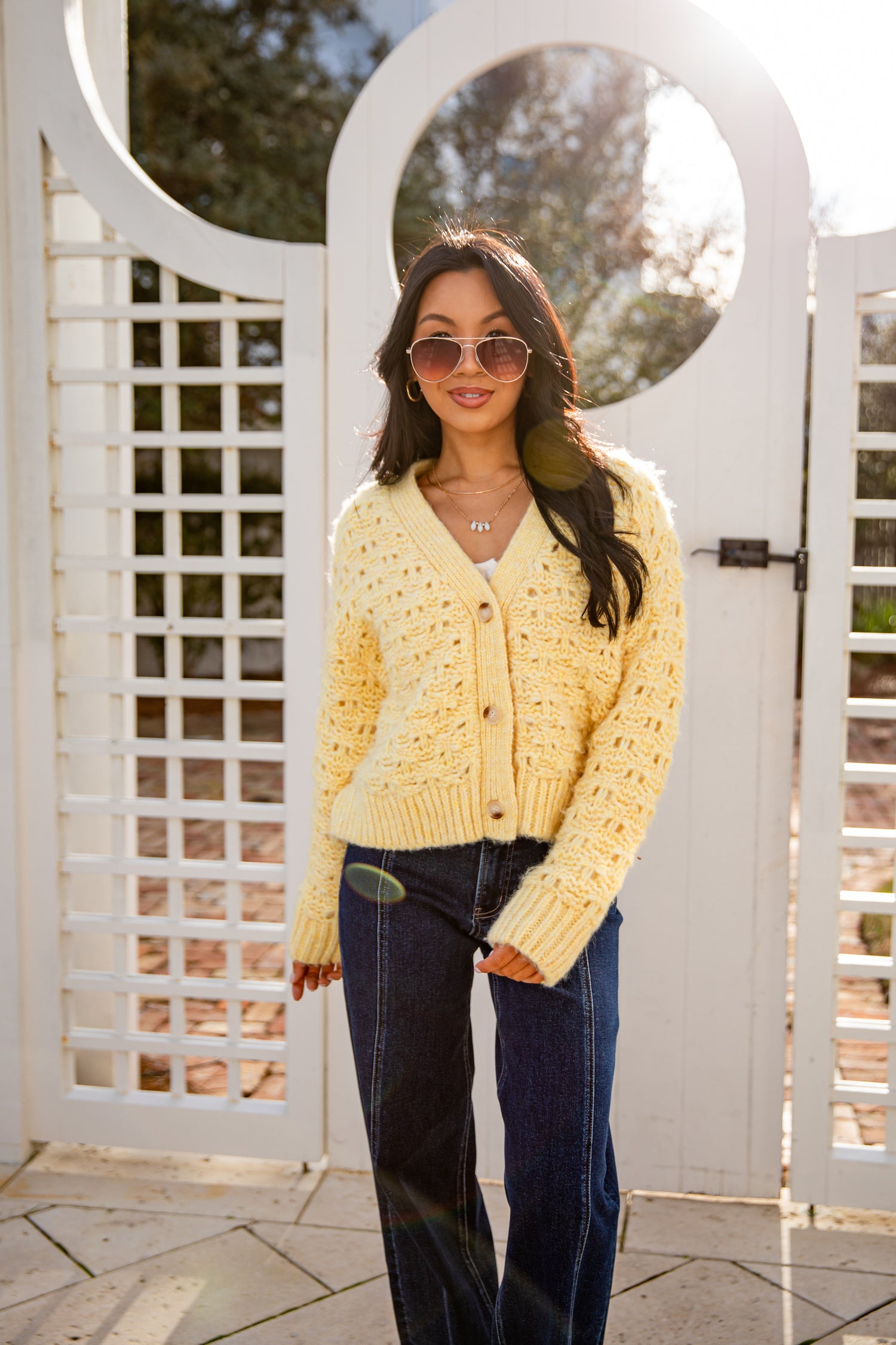 Woman wearing a yellow cardigan and sunglasses standing in front of a white lattice gate.